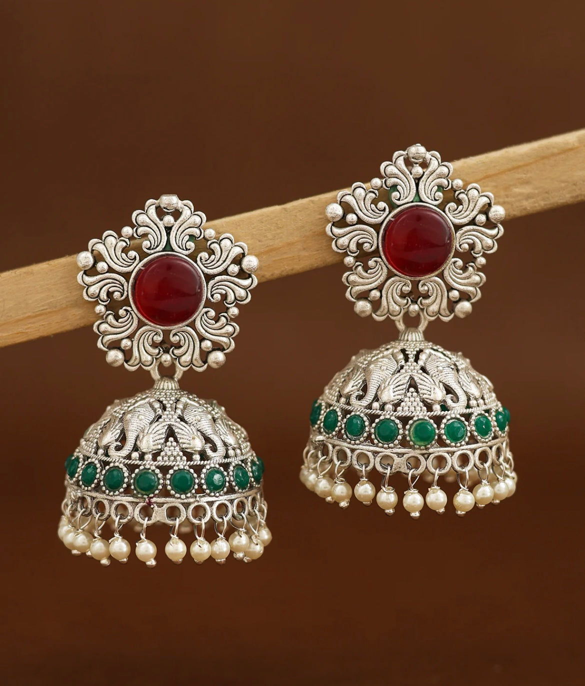 Ornate silver earrings with red and green stones on a brown background